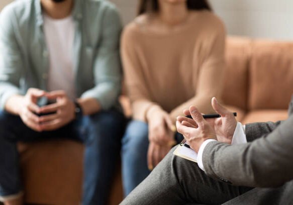 Help Concept. Closeup Cropped View Of Male Counselor Therapist Or Consultant In Suit Talking With Couple Sitting In Office, Giving Professional Advice. Selective Focus On Hands With Clipboard And Pen