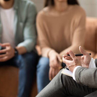 Help Concept. Closeup Cropped View Of Male Counselor Therapist Or Consultant In Suit Talking With Couple Sitting In Office, Giving Professional Advice. Selective Focus On Hands With Clipboard And Pen